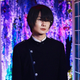 Kamiki is dressed as his character, standing in front of an open door with purple flowers in the background.